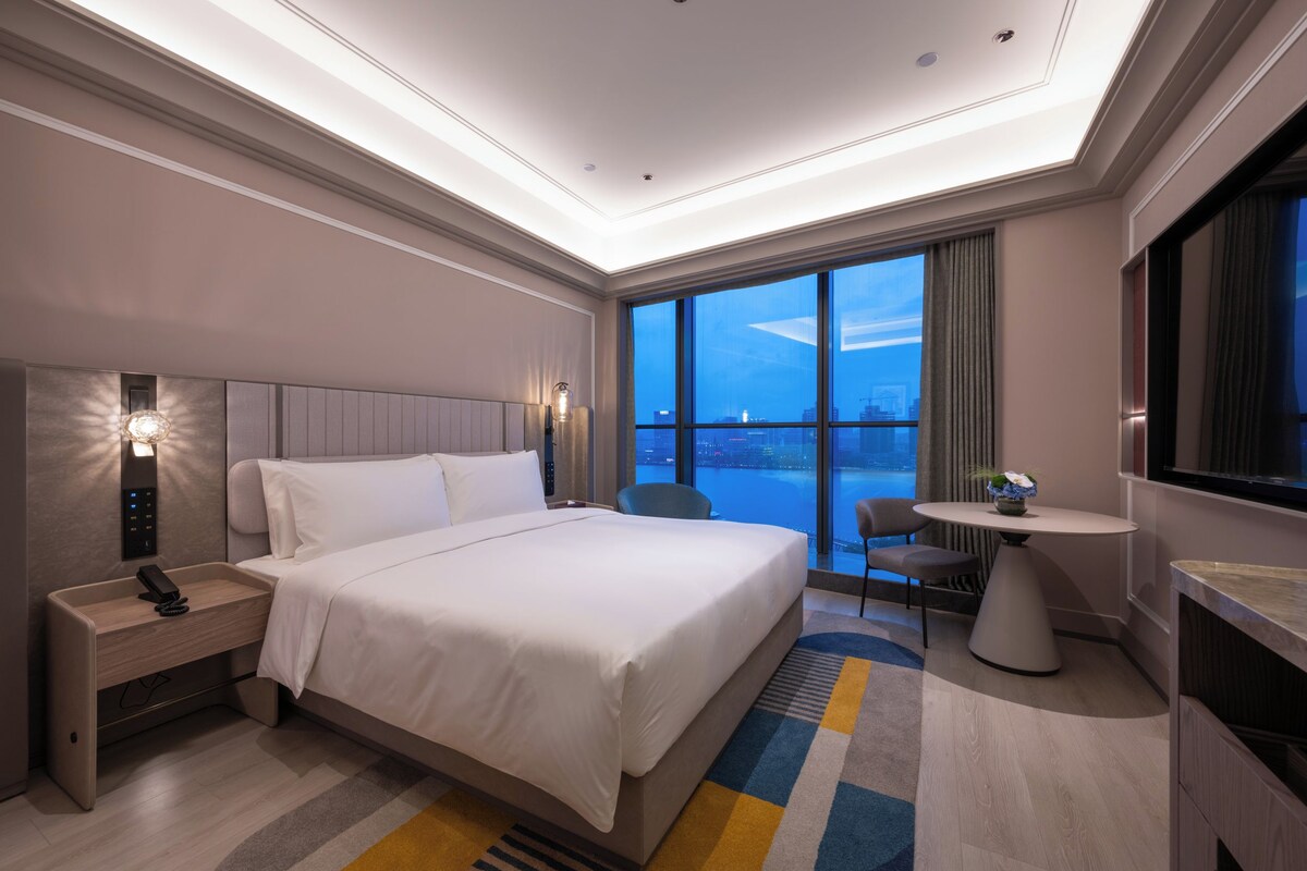 A hotel room with a large bed, bedside controls, a desk with a phone, a chair, a small table with a vase, and a large window offering a city view.