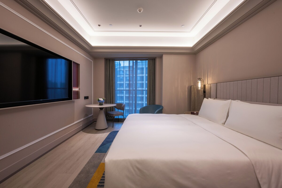 A hotel room with a large bed, a flat-screen TV, a small table with a vase, and a view of the city through the window.