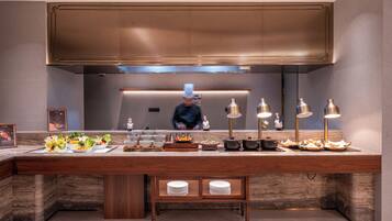 A modern kitchen with a chef preparing food, a buffet setup with various dishes, and a wooden counter with plates.
