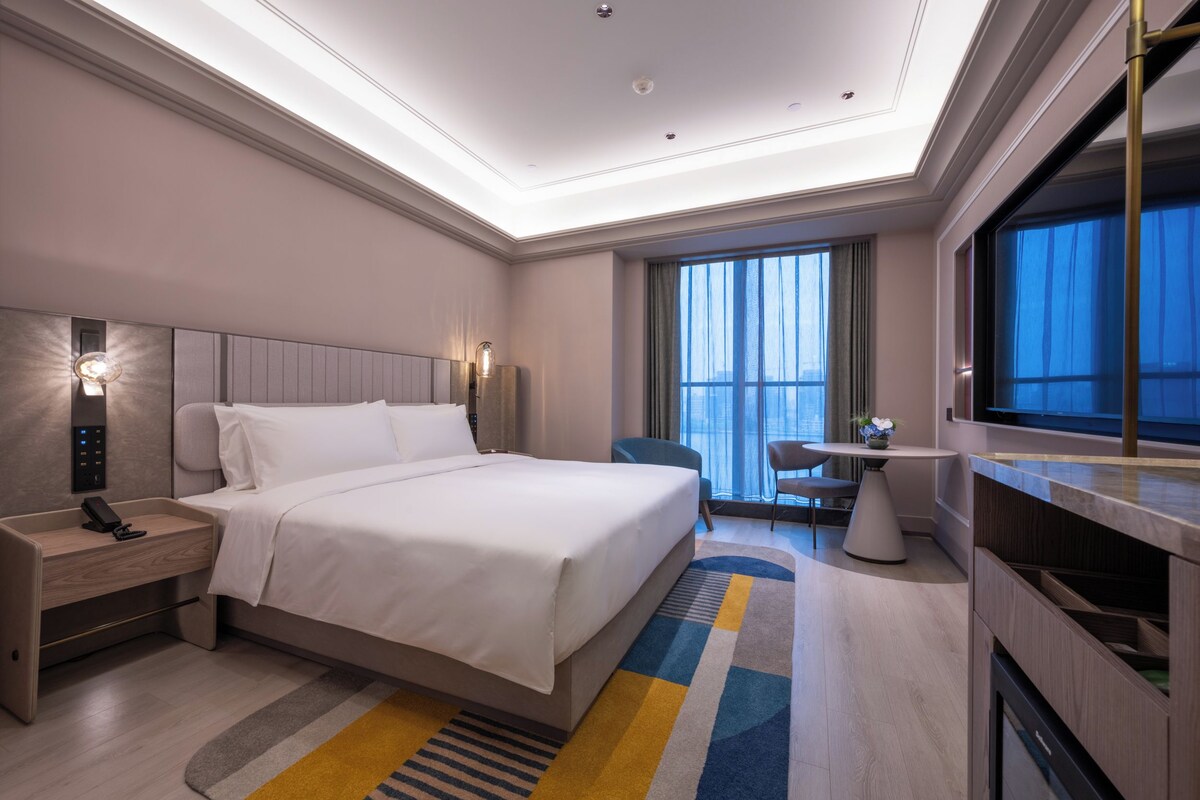 A modern hotel room with a large bed, bedside table with a phone, a desk with a chair, a window with curtains, and a view of the city.