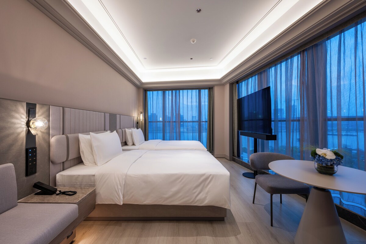 A modern hotel room with a large bed, bedside tables, a TV, a seating area, and large windows with curtains.