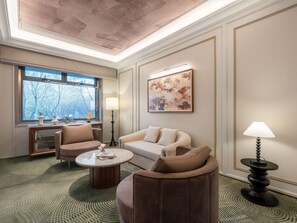 Un salon moderne avec un canapé, des fauteuils, une table basse et une lampe. Un tableau orne le mur et une fenêtre offre une vue sur les arbres.