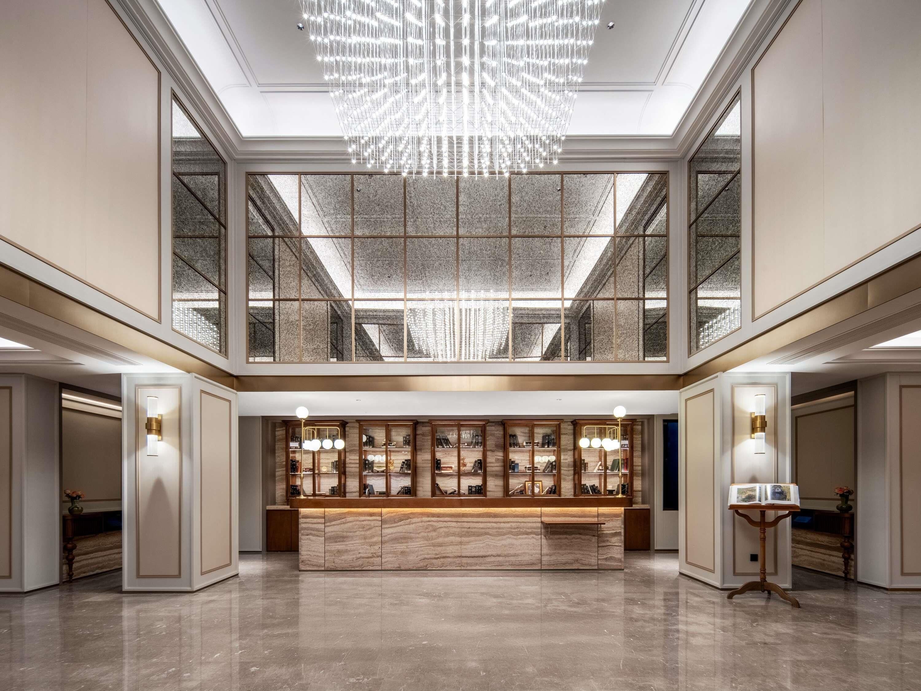 A spacious lobby with a marble reception desk, a large chandelier, and a high ceiling with skylights.
