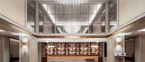 A spacious lobby with a marble reception desk, a large chandelier, and a high ceiling with skylights.