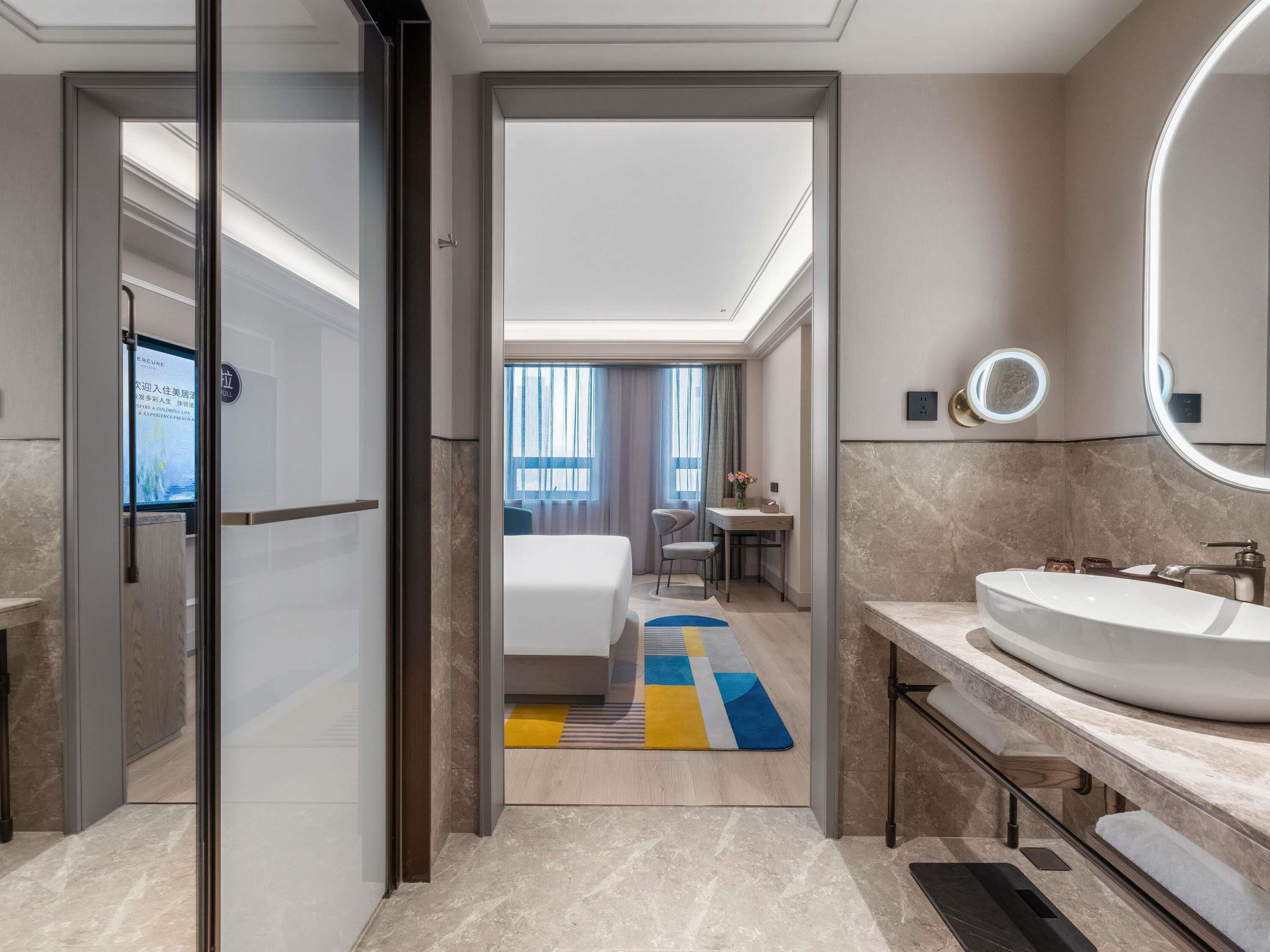 A modern hotel bathroom with a large sink, a mirror, and a view into the bedroom.