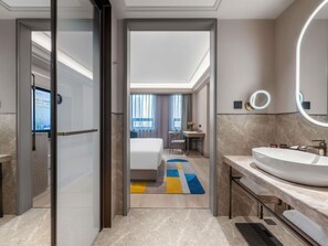 A modern hotel bathroom with a large sink, a mirror, and a view into the bedroom.