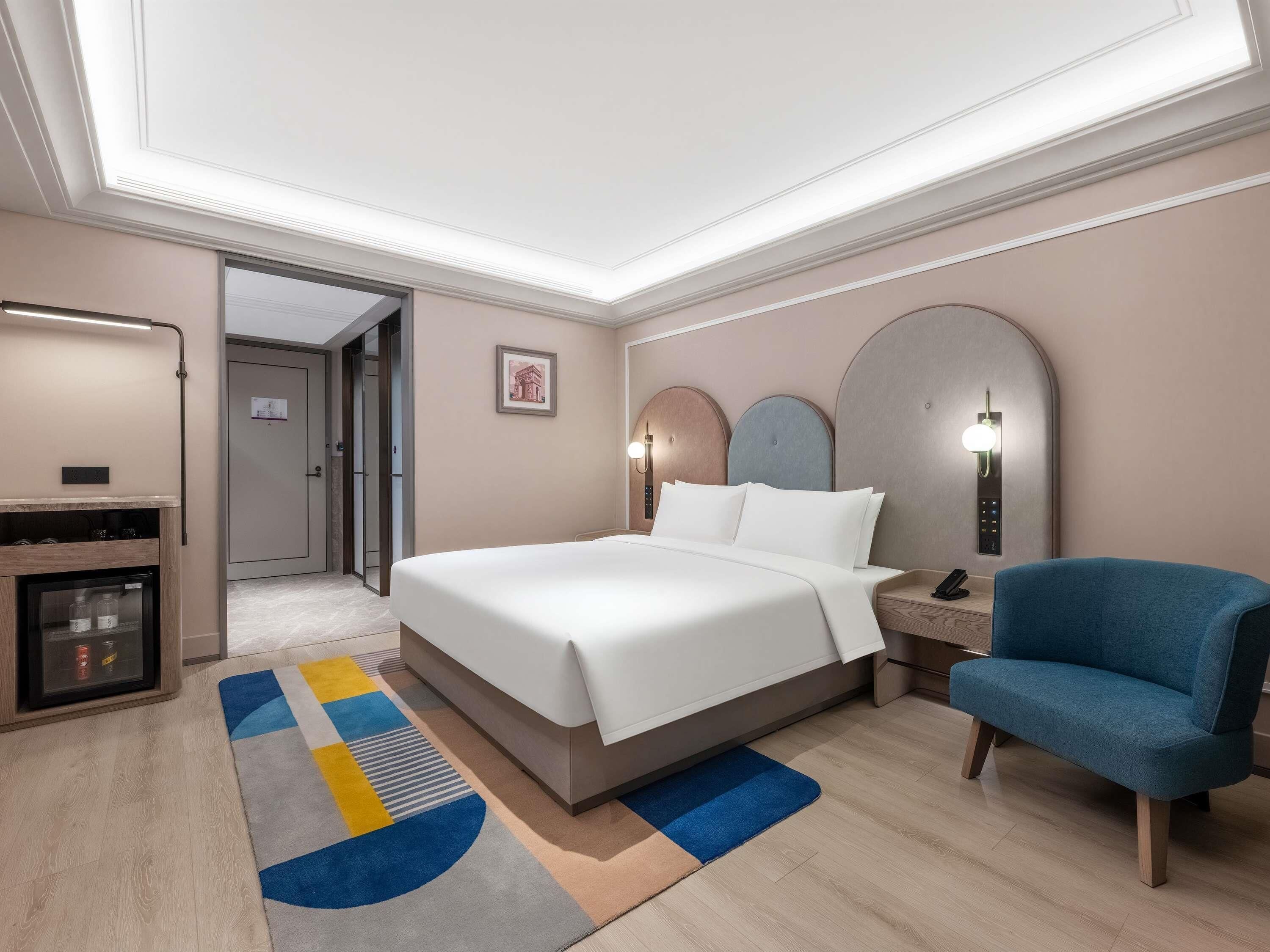 A hotel room with a bed, a desk, a chair, a minibar, and a door leading to another room.