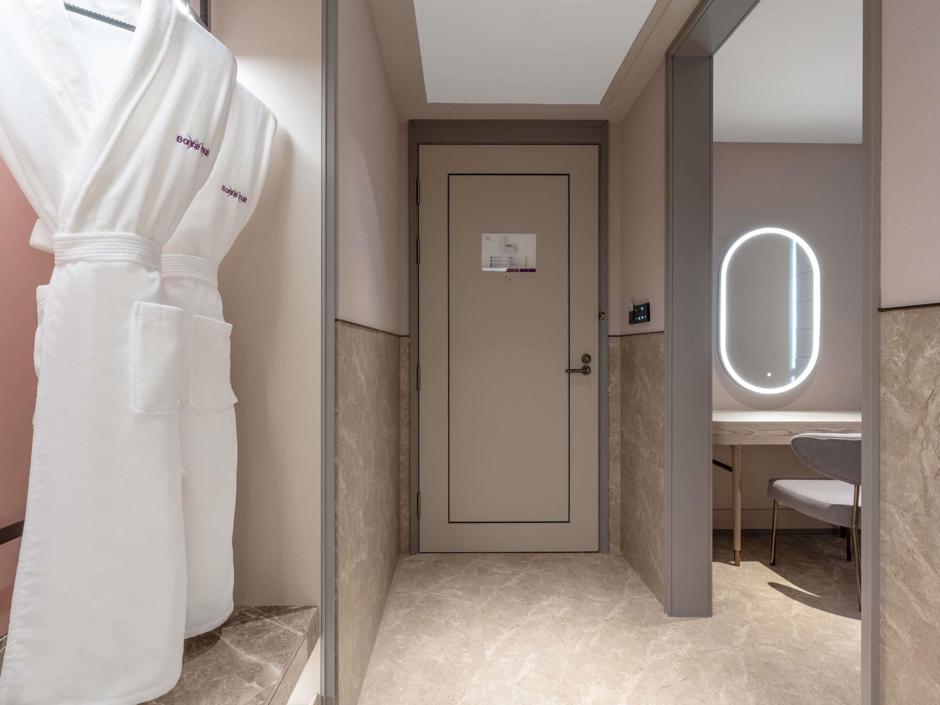 A hotel bathroom with a door, a robe, a mirror with lights, and a sink.