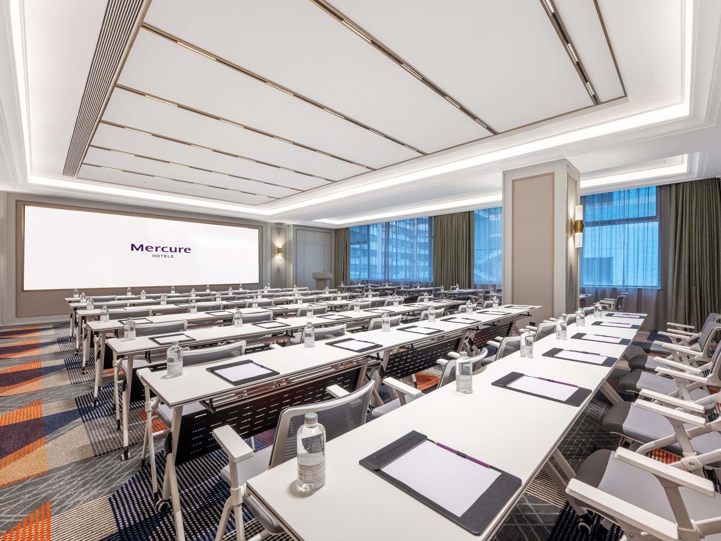 Un salón de conferencias con filas de mesas, sillas y una gran pantalla mostrando 'Mercure Hotels'.