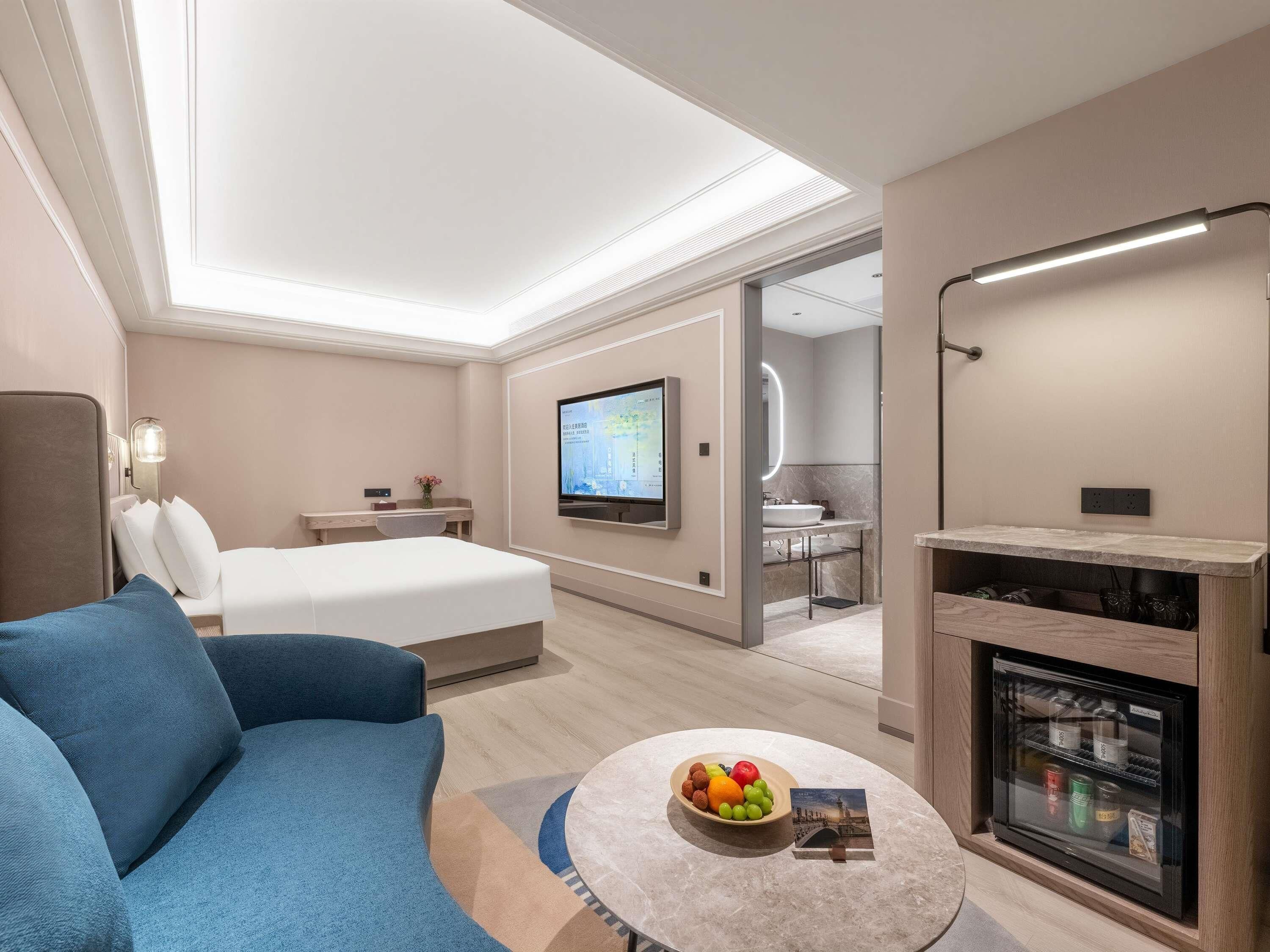 A modern hotel room with a bed, a television, a small desk, a sofa, a coffee table with fruit and a magazine, and a fireplace.