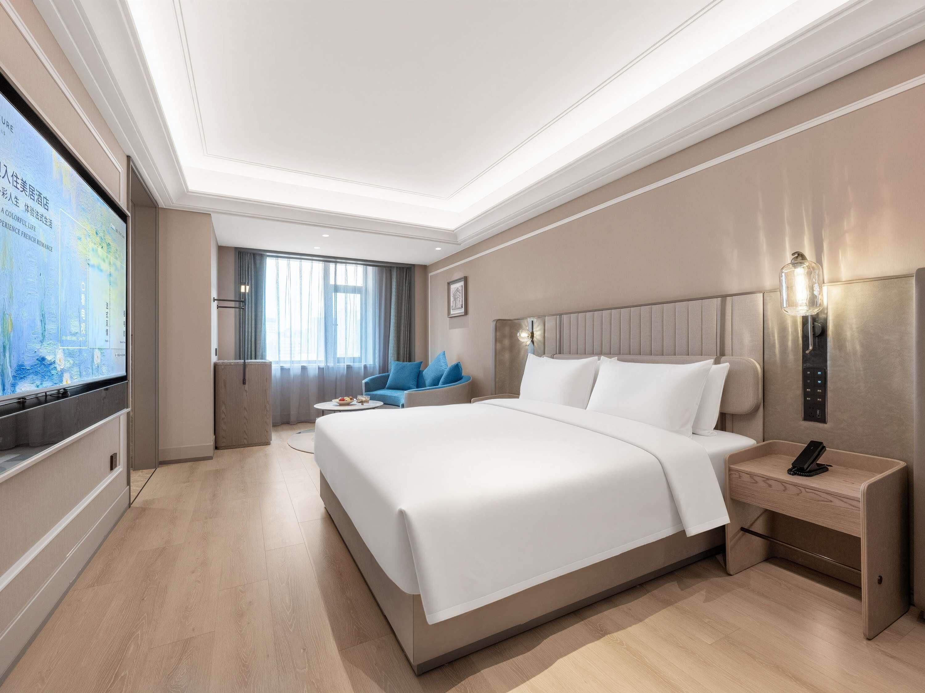 A hotel room with a large bed, a bedside table with a lamp, a TV mounted on the wall, and a seating area with a chair and a small table.