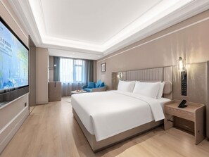 Ein Hotelzimmer mit einem großen Bett, einem Nachttisch inklusive Lampe, einem an der Wand montierten Fernseher und einer Sitzecke mit Sessel und kleinem Tisch.