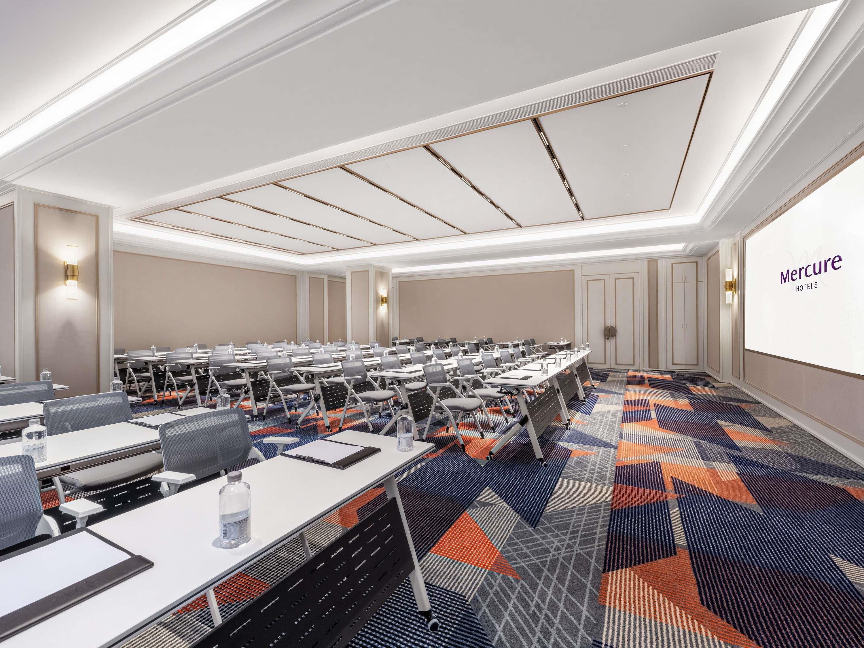 A conference room with rows of tables and chairs, a patterned carpet, and a large screen displaying 'Mercure Hotels'.