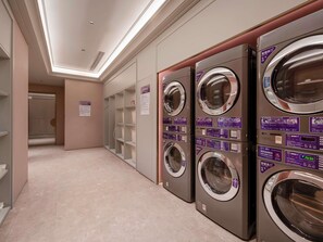 A row of wall-mounted washers with control panels, a tiled floor, and a door leading to another room.