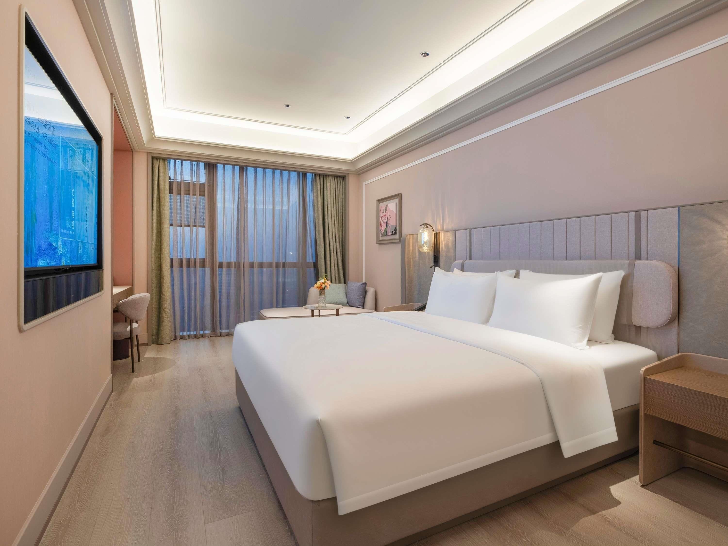 Una habitación de hotel moderna con una cama grande, un escritorio y vistas de la ciudad a través de cortinas translúcidas.