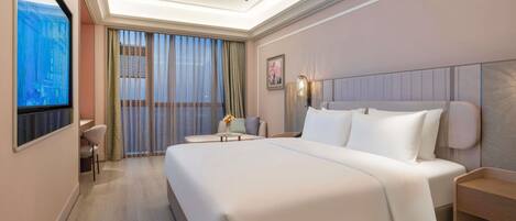A modern hotel room with a large bed, a desk, and a view of the city through sheer curtains.