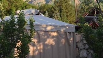A tented area with a stone wall, surrounded by trees and a mountain backdrop.