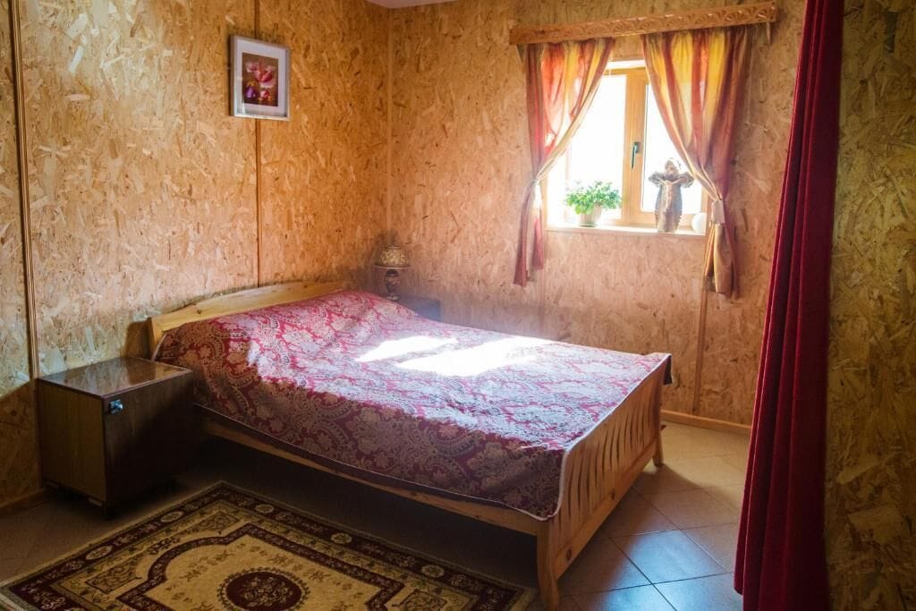 Un dormitorio con cama, mesita de noche, ventana con cortinas e imagen colgada en la pared.