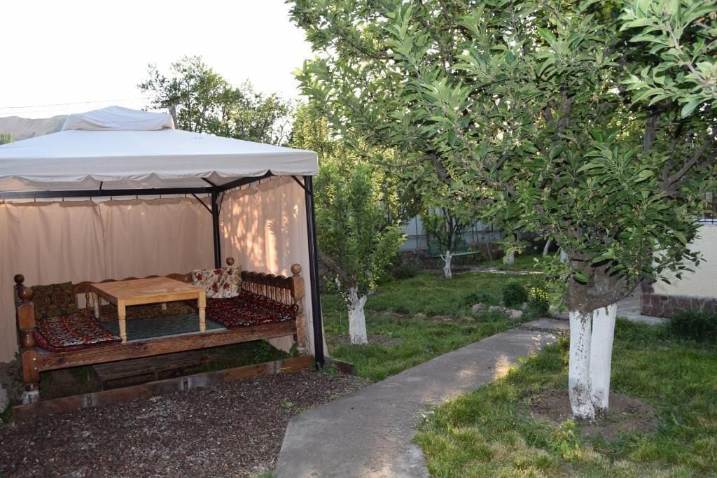 Un espacio de descanso cubierto al aire libre, con banco y mesa, rodeado de árboles y un sendero en el jardín.