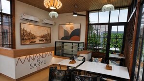A restaurant with a wooden ceiling, black chairs, and tables. There are paintings on the walls and a large window with a view.