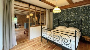 A bedroom with a wooden floor, a bed with a metal frame, a window with curtains, and a hanging light fixture.