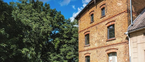 A brick building with multiple windows and a door, surrounded by lush greenery and a clear sky.