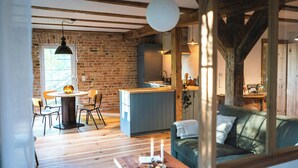 A rustic interior with exposed wooden beams, a brick wall, a dining area with a round table and chairs, a kitchenette, and a sofa with a view.