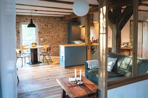 A rustic interior with exposed wooden beams, a brick wall, a dining area with a round table and chairs, a kitchenette, and a sofa with a view.