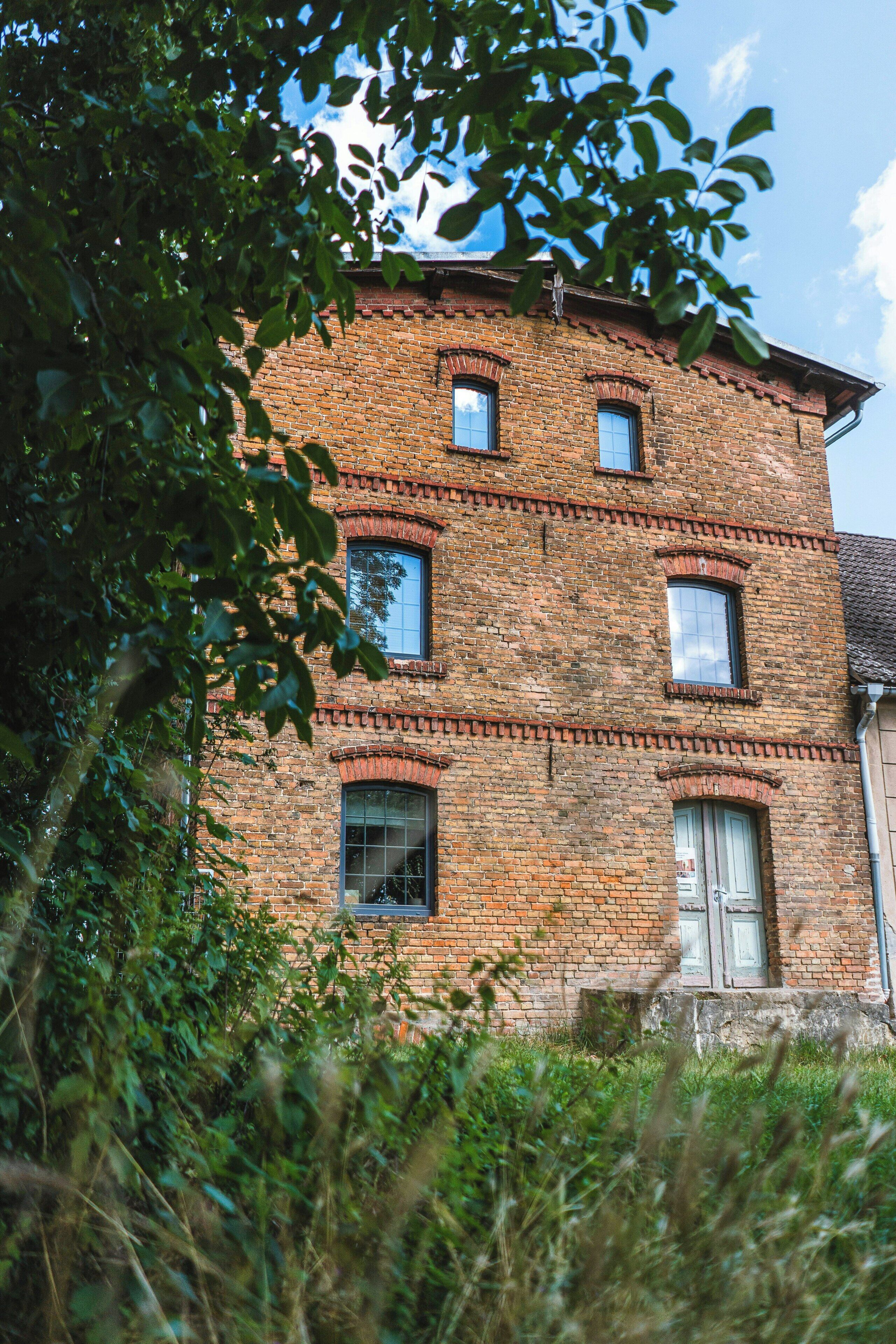 A brick building with multiple windows and a door, surrounded by greenery.
