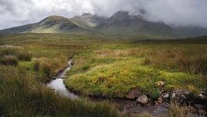 A natural landscape with a stream, grassy fields, and mountains in the background.