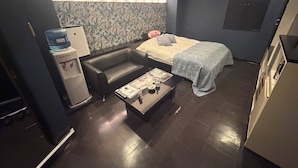 A compact hotel room with a sofa, a bed, a small table with a magazine, a water dispenser, and a wall with a floral pattern. - HOTEL PALETTE GOLD (Kurashiki)