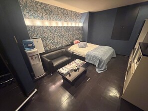 A compact hotel room with a sofa, a bed, a small table with a magazine, a water dispenser, and a wall with a floral pattern.