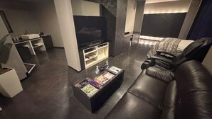 A modern living room with a black leather sofa, a flat-screen TV, a coffee table with a menu, and a small kitchen area. - HOTEL PALETTE GOLD (Kurashiki)