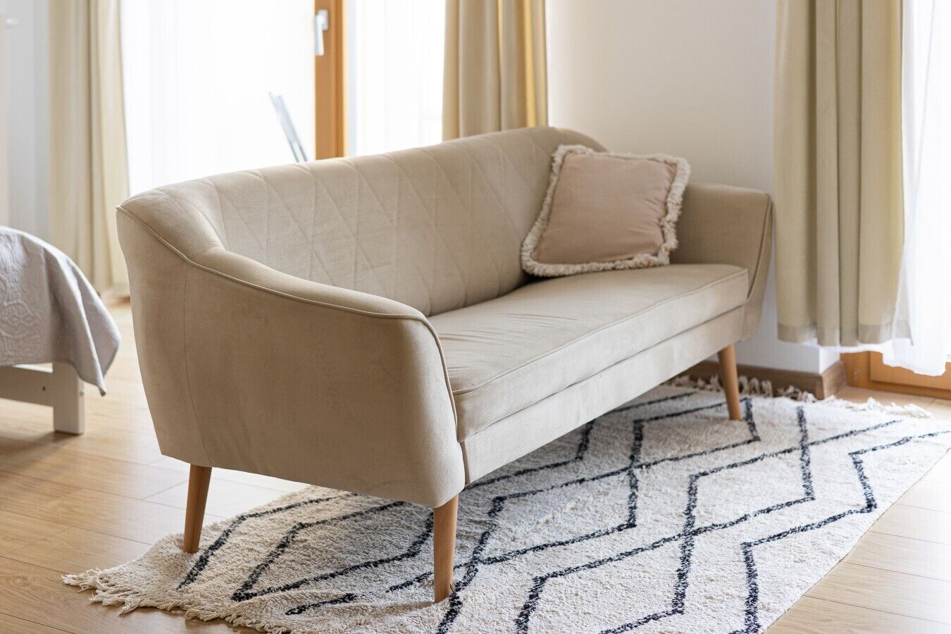 Ein beiges Sofa mit Holzbeinen, ein passendes Kissen und ein gemustertes Teppich auf Holzdielen.