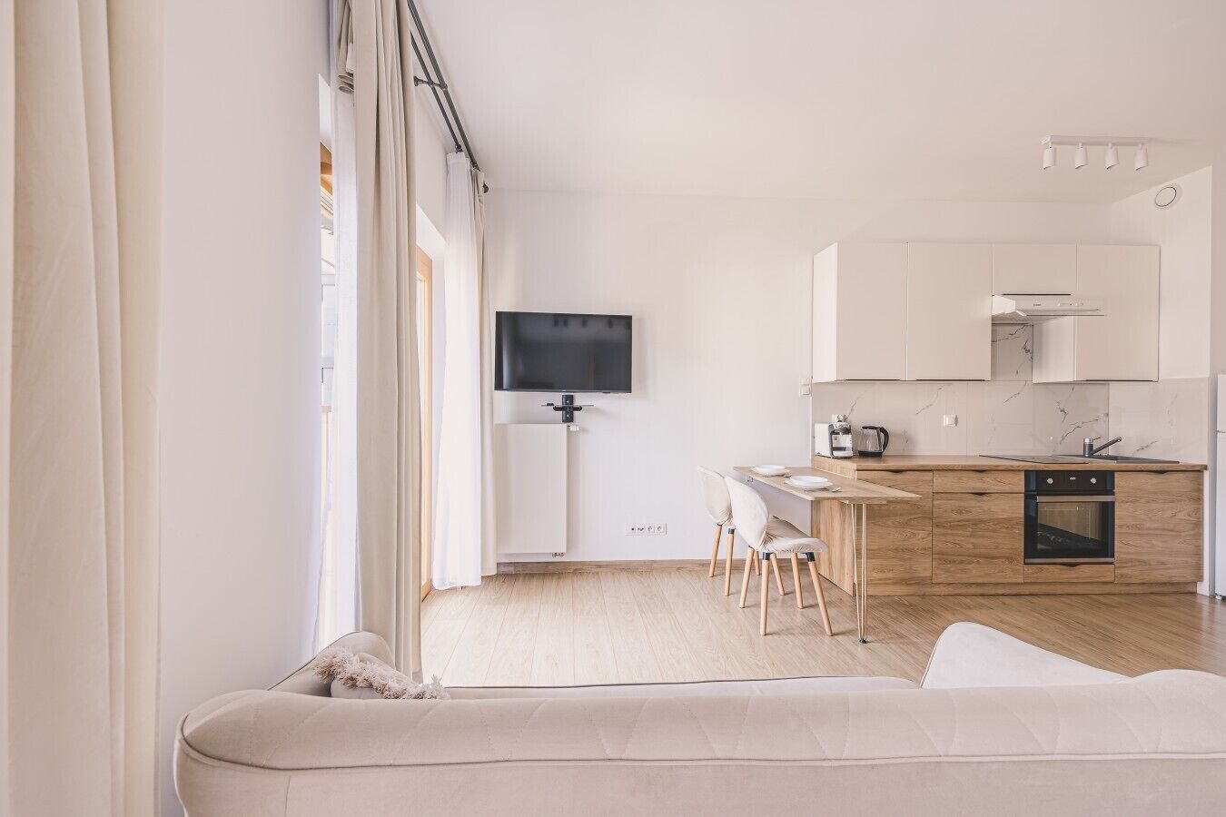 Ein modernes Wohnzimmer mit einer Couch, einem Essbereich mit Stühlen, einer Küche mit Schränken und Geräten sowie einem an der Wand befestigten Fernseher.