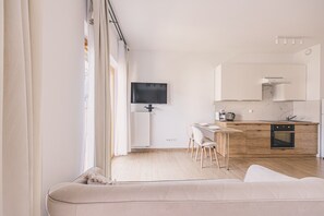 Una sala de estar moderna con un sofá, un comedor con sillas, una cocina con armarios y electrodomésticos, y un televisor montado en la pared.