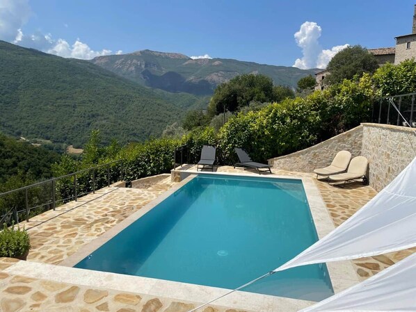 Piscina rectangular con plataforma de piedra, rodeada de sillones de descanso y una pérgola blanca, con vistas a un paisaje montañoso.