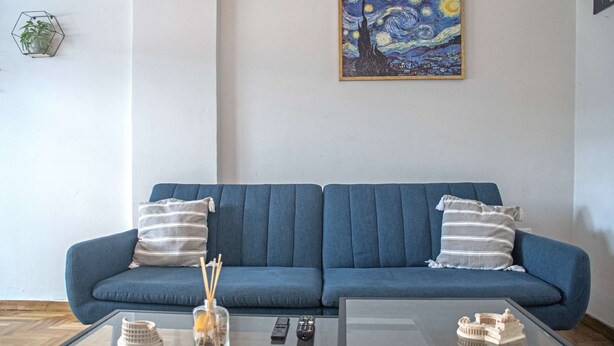 Una sala de estar con un sofá azul, dos almohadas a rayas, una mesa de centro de vidrio con objetos decorativos y un cuadro de una escena nocturna en la pared.