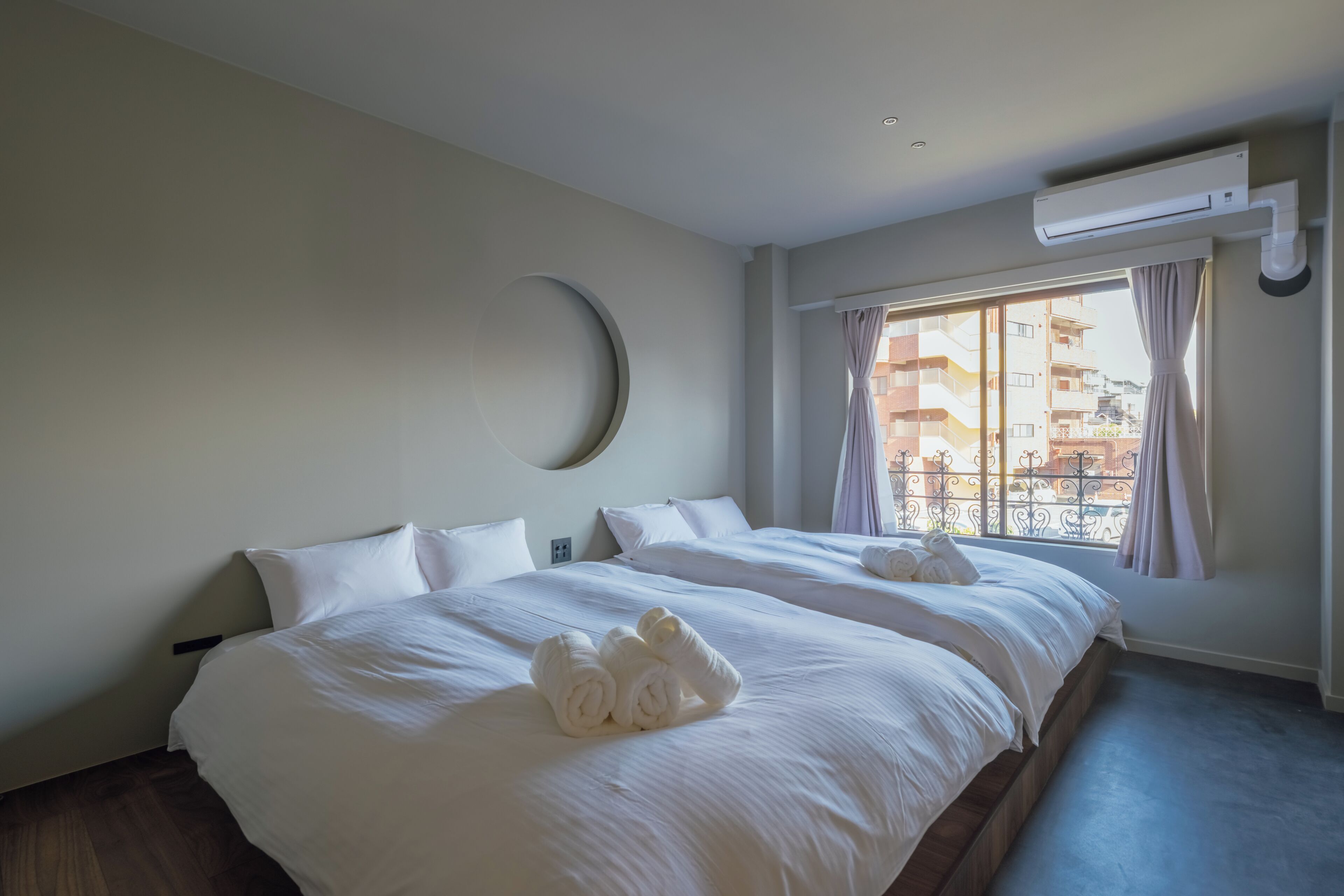 Quarto de hotel com duas camas, uma janela grande com cortinas, um aparelho de ar condicionado e uma decoração em forma de crescente na parede.