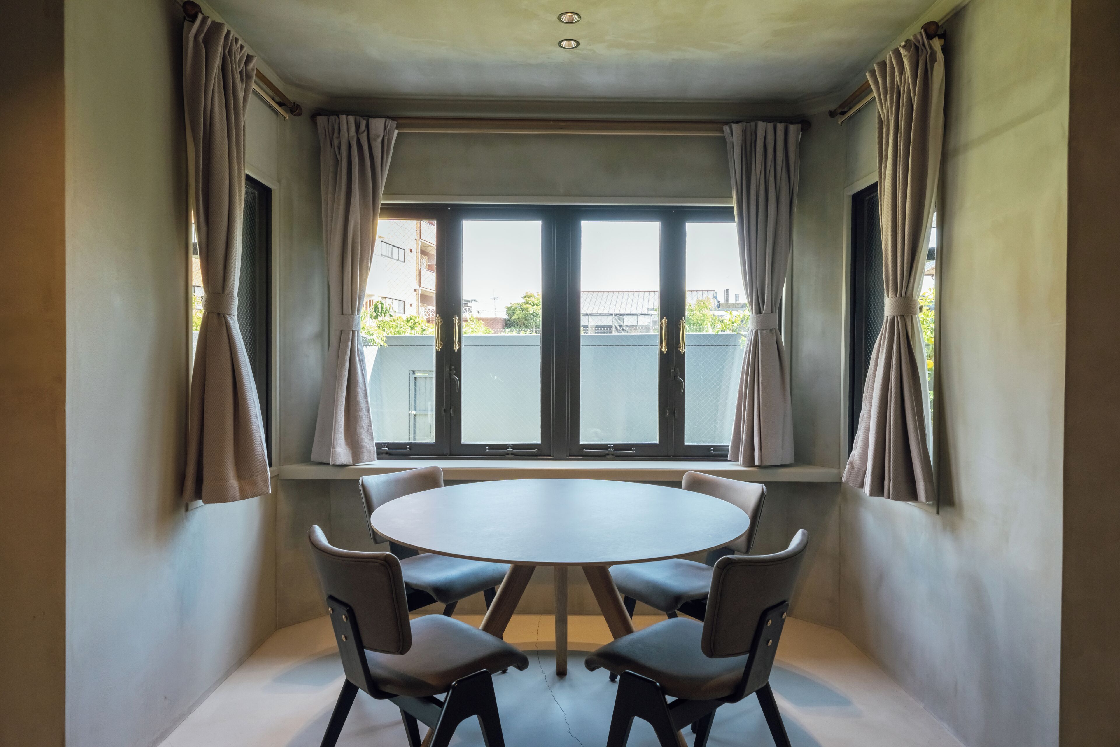Una habitación con una mesa redonda y cuatro sillas, un ventanal con cortinas y vistas a edificios.