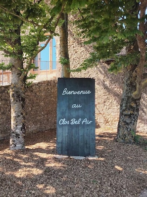 Uma placa de madeira com o texto 'Bienvenue au Clos Bel Air' em um cenário natural com árvores e um muro de pedra.