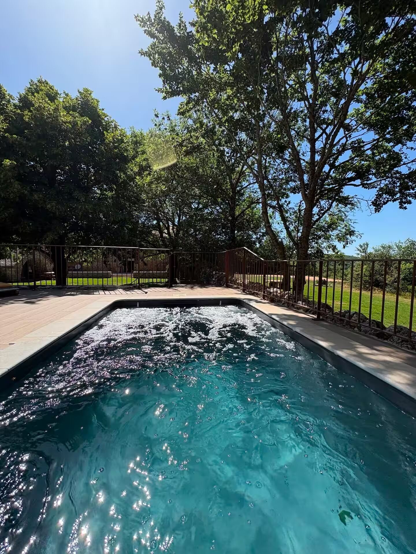 A swimming pool with a clear blue water surface, surrounded by a paved deck and a metal fence.