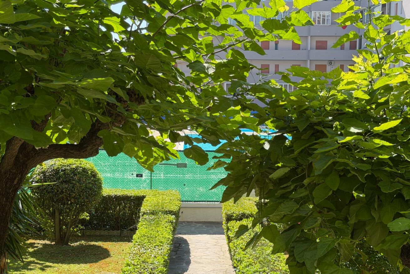 Um caminho ladeado por sebes e árvores bem aparadas, que leva a um edifício com telhado verde.