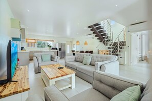 A modern living room with a sofa, coffee table, and TV. There is a kitchen in the background and a staircase leading to an upper level. - Modern Two Villas • 6BR • 2 Private Pools • Aruba Escape (Noord)