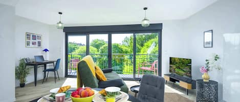 A modern living room with a dining area, a sofa, a TV, and a balcony with a view of greenery.