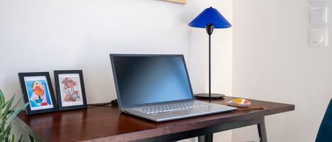 A laptop on a desk with a blue lamp, two framed artworks, and a small orange object.