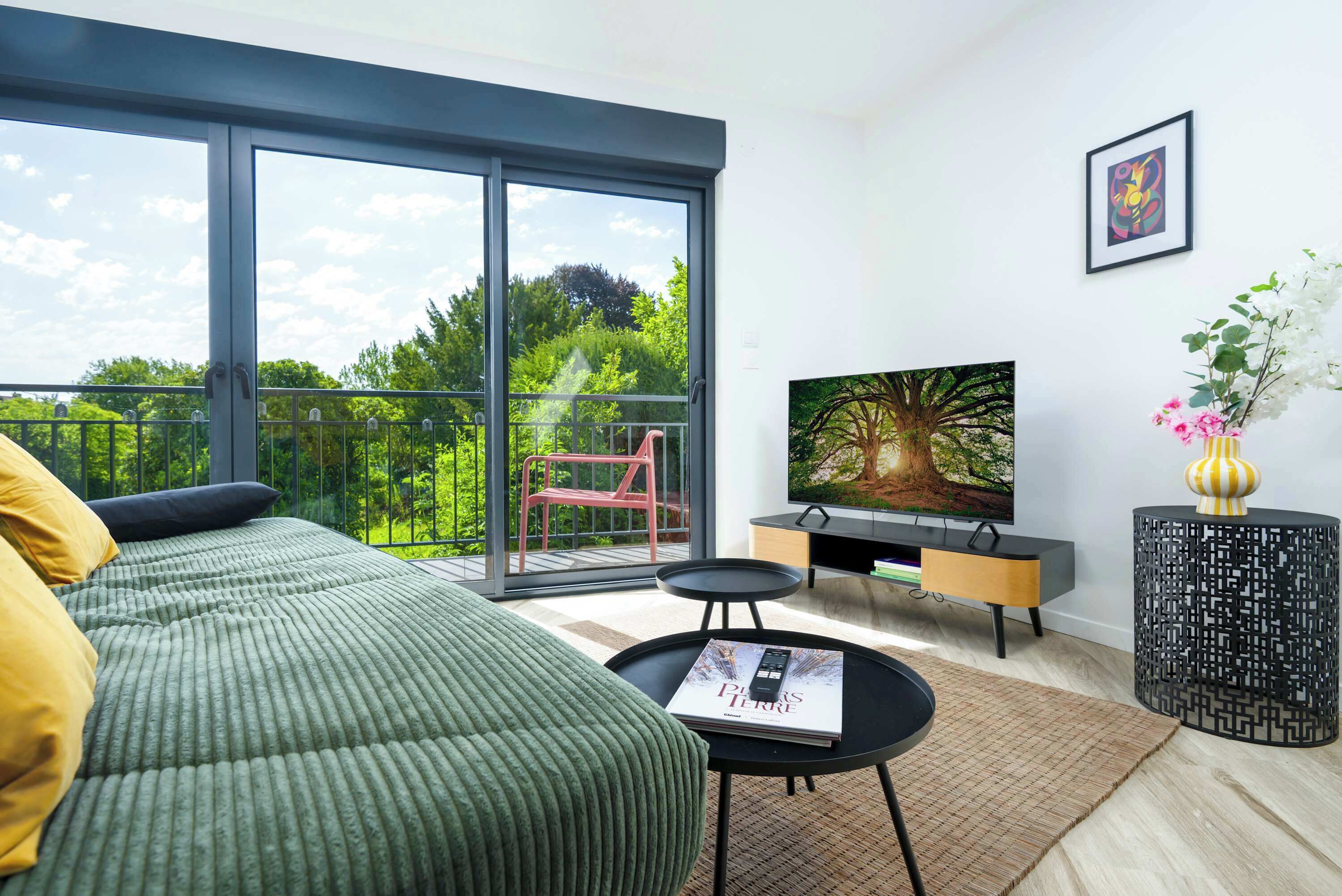 A modern bedroom with a large sliding glass door leading to a balcony, a bed with green bedding, a television on a stand, and a small table with a remote control.