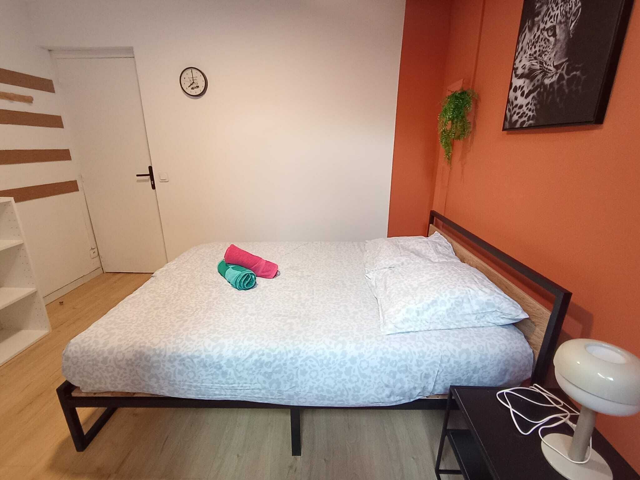 Un dormitori amb un llit, una taula de nit, un rellotge de paret i una fotografia emmarcada a la paret.