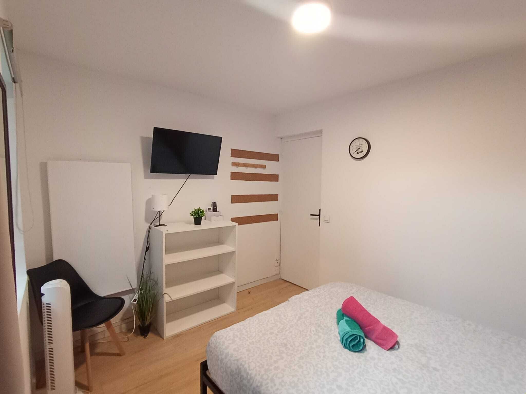 A bedroom with a bed, a TV mounted on the wall, a desk with a chair, and a plant.