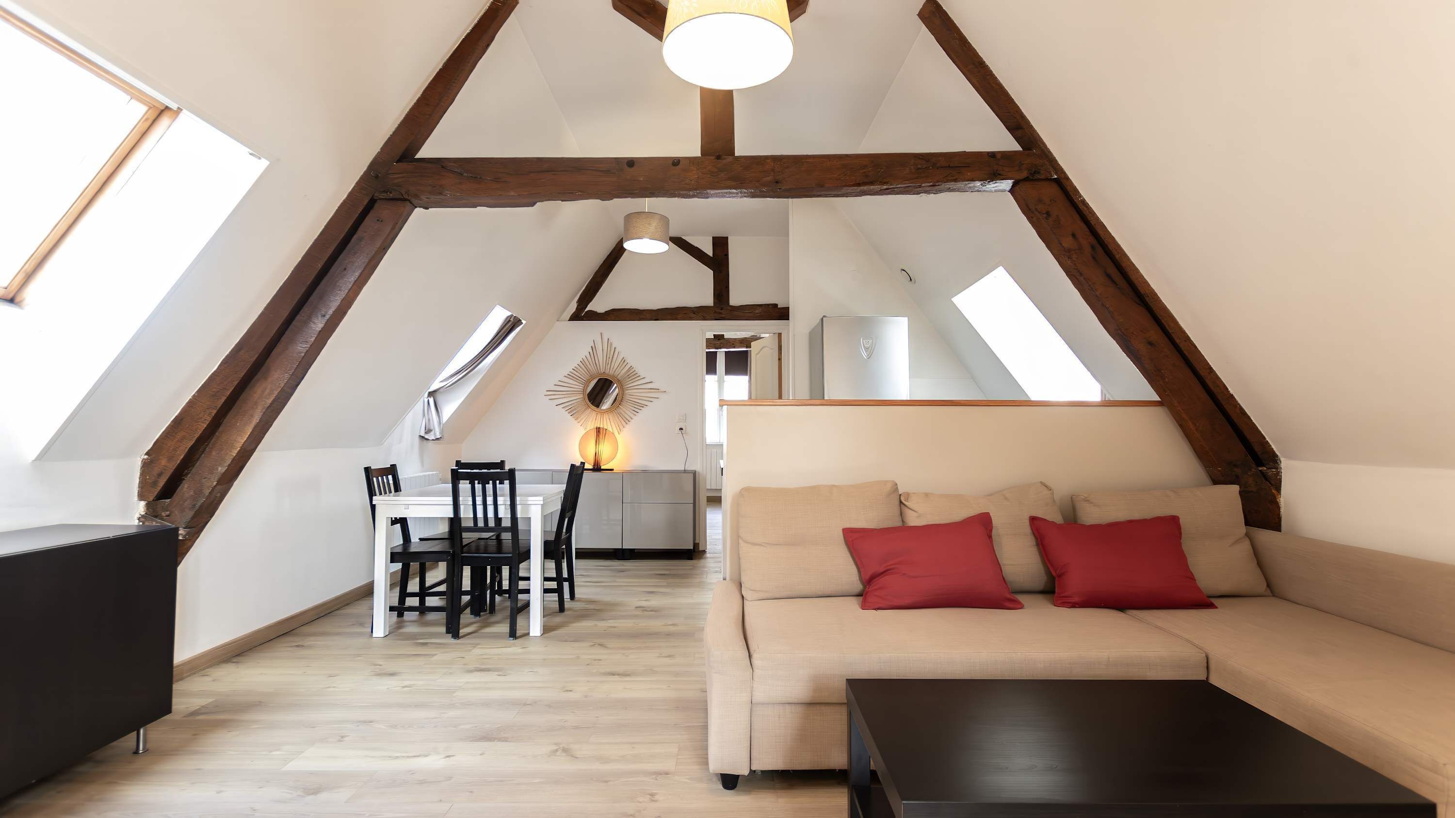 A loft-style room with a sloped ceiling, wooden beams, a sofa with red cushions, a dining table with chairs, and a lamp.